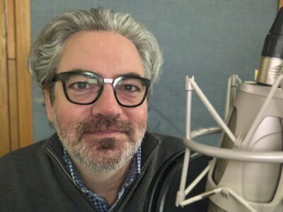 Jonathan K. Riggs in the recording studio behind a microphone. Riggs is wearing glasses and a grey sweater over a blue checkered shirt.