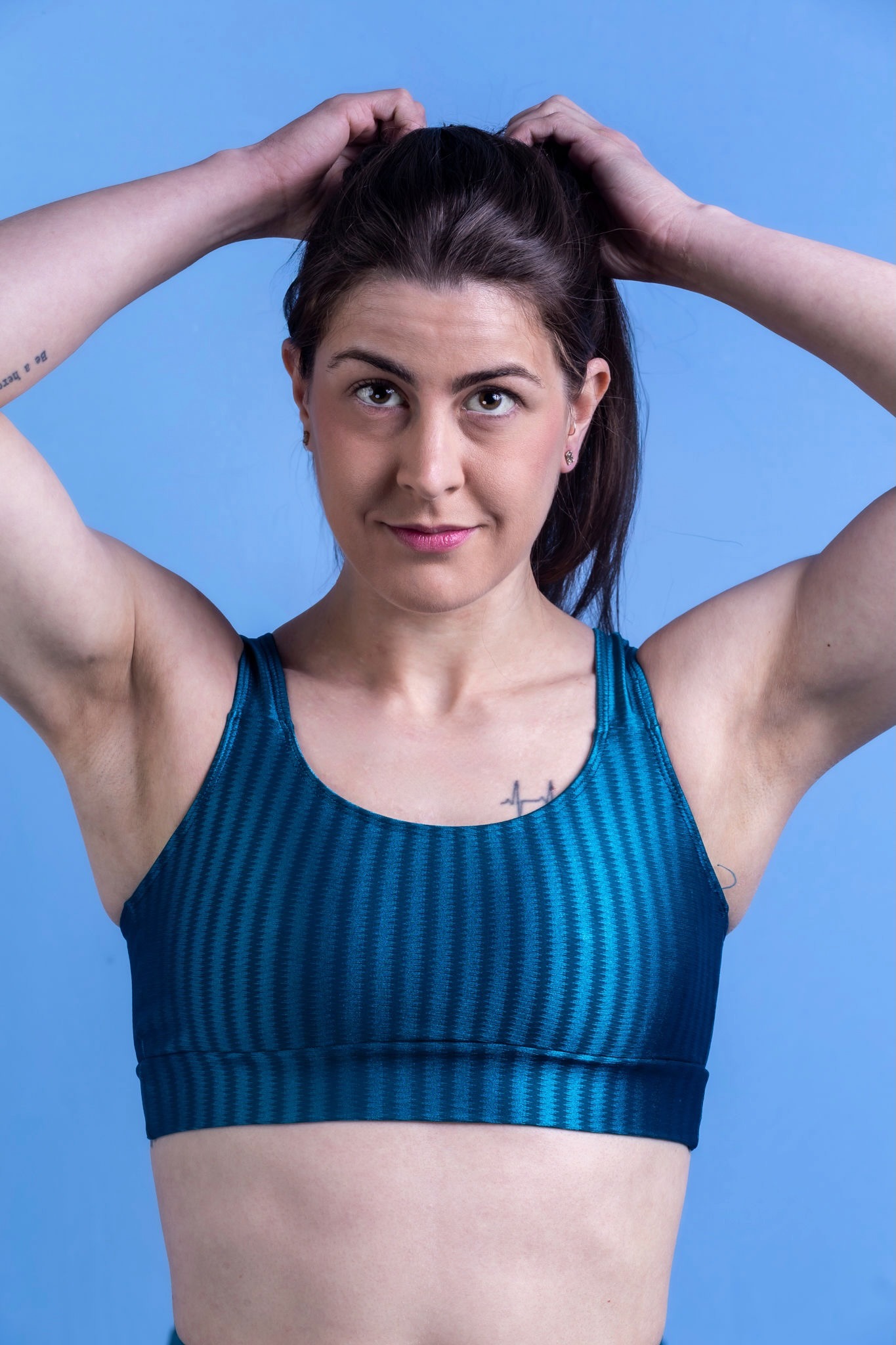 Jules Franchetto shows her physique in a blue sports bra and arms raised to adjust her ponytail. She has brown eyes, stud earrings, and lipstick.