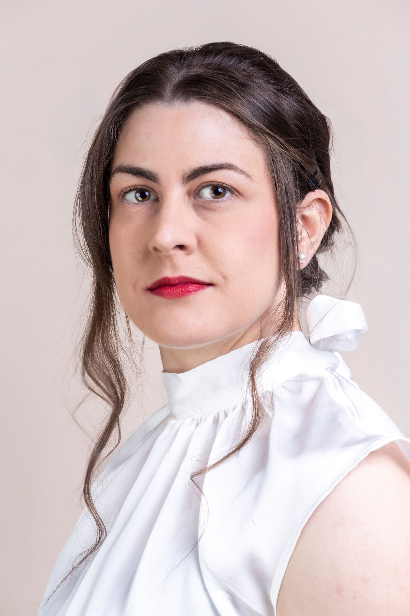 This is a professional headshot photograph of Jules Franchetto. Her brunette hair is elegantly fixed with bobby pins and a couple of curled strands framing her face. She has brown eyes and is wearing pearl earrings and red lipstick.  They are wearing a white silk blouse with a bow in the back. 