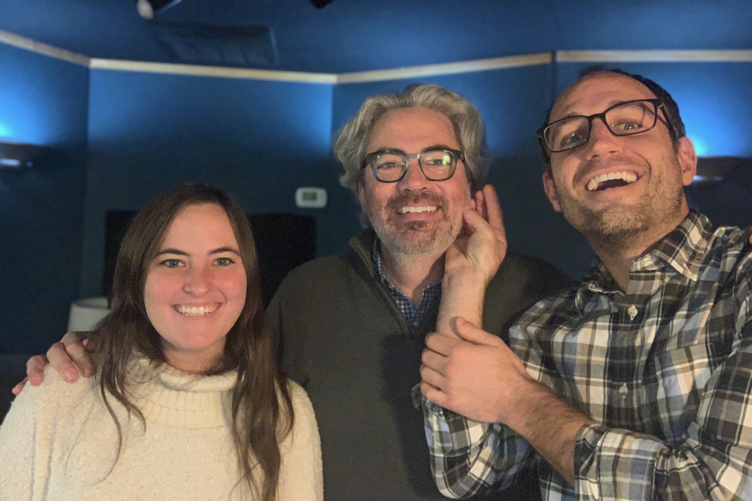 Jonathan K. Riggs in the control room of a recording studio with Sydney Mathieu (Left) with long brunette hair and a cream-colored sweater, and Craig Hinkle (Right) with glasses and a plaid shirt. Jonathan is in glasses and a grey sweater over a blue checkered shirt.