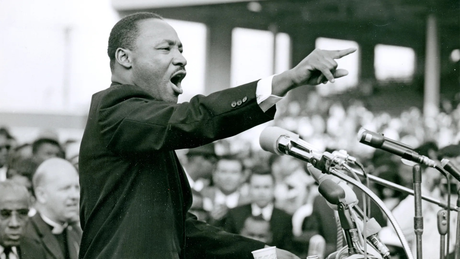 A black and white photograph of Dr. Martin Luther King, Jr. behind a podium with several microphones.